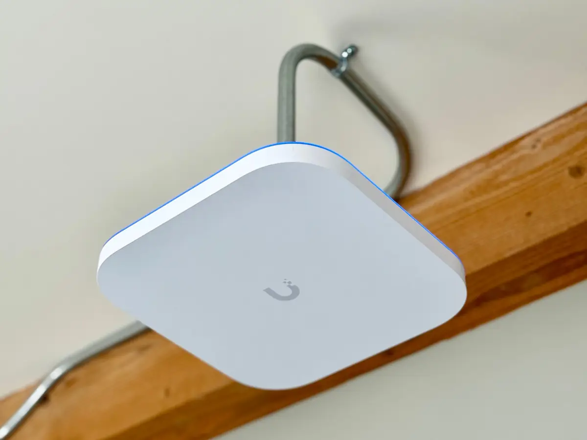 White UniFi access point on a conduit hanger below a wood beam