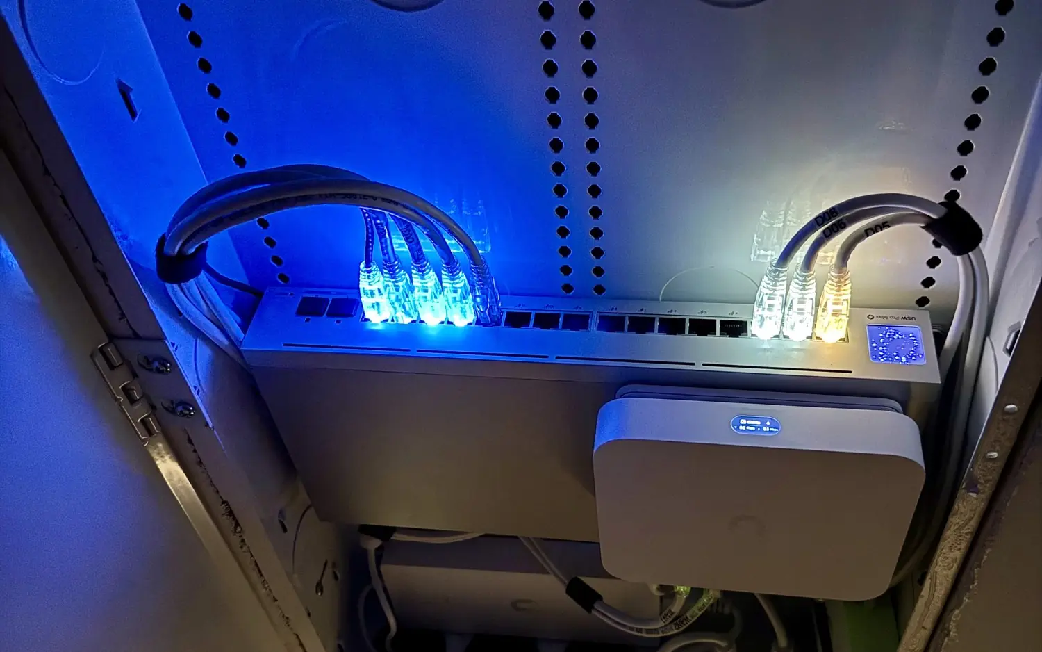 UniFi switch and gateway in a wall enclosure lit blue, seen from below